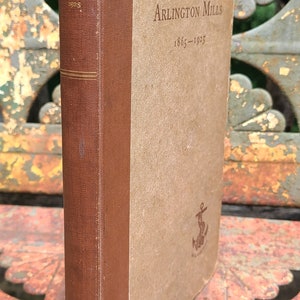 May include: A brown book with a tan spine. The book is titled "Arlington Mills 1865-1925" and has a small anchor symbol on the front cover.
