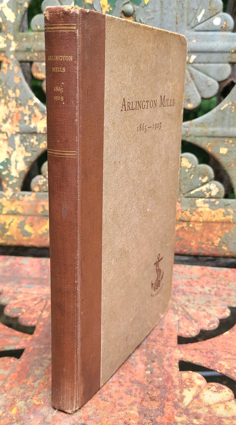 May include: A brown book with a tan spine. The book is titled "Arlington Mills 1865-1925" and has a small anchor symbol on the front cover.