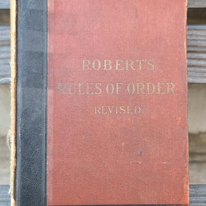 May include: A vintage red book with black accents and gold lettering. The title is "Robert's Rules of Order: Revised".