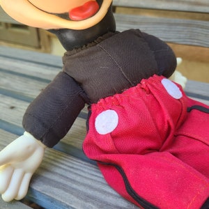 May include: A vintage plush Mickey Mouse toy with black fabric for the torso and red fabric for the trousers. The trousers have white circles on the sides and black trim.