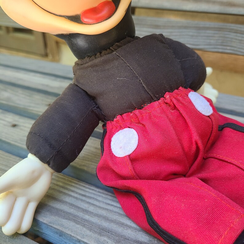 May include: A vintage plush Mickey Mouse toy with black fabric for the torso and red fabric for the trousers. The trousers have white circles on the sides and black trim.
