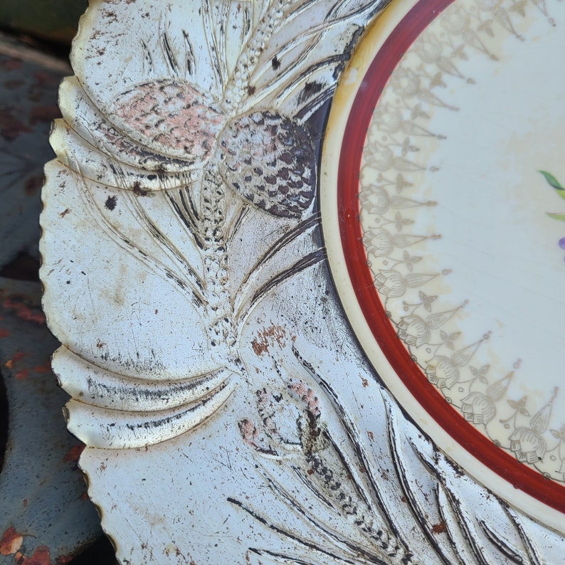 May include: A white ceramic plate with a red and gold rim. The plate has a floral design and is decorated with a raised, embossed design of pine branches and cones.