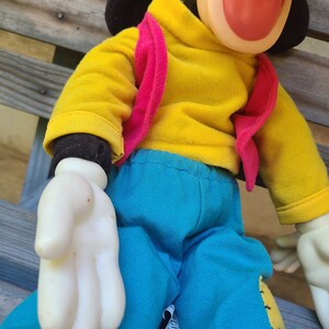 May include: A plush toy with a black head, yellow shirt, pink vest, and blue trousers. The toy has a large, red, open mouth.