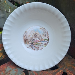 May include: White ceramic bowl with a scalloped rim and a colorful scene of a wintery village with a stream and trees.