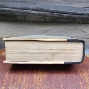 May include: A thick book with aged, cream-colored pages. The book's spine is dark blue with a black edge, and the bottom edge has a red accent. The book is resting on a rusty, textured surface.