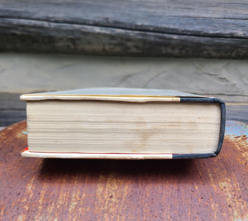 May include: A thick book with aged, cream-colored pages. The book's spine is dark blue with a black edge, and the bottom edge has a red accent. The book is resting on a rusty, textured surface.