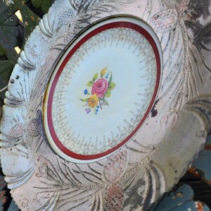 May include: A white ceramic plate with a raised, ornate design. The center of the plate features a floral motif with pink, yellow, and blue flowers. The plate has a red and gold rim.