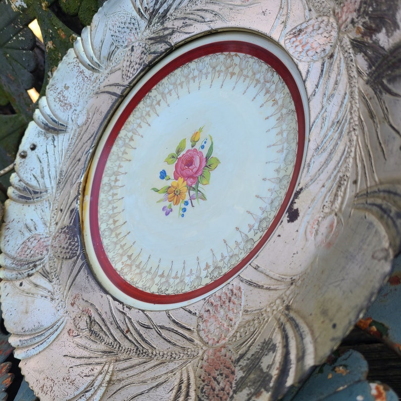 May include: A white ceramic plate with a raised, ornate design. The center of the plate features a floral motif with pink, yellow, and blue flowers. The plate has a red and gold rim.