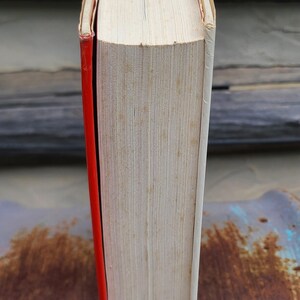 May include: A hardcover book with a red spine and white pages. The top edge of the book is visible, showing the pages and a black and red detail. The book's cover is white.