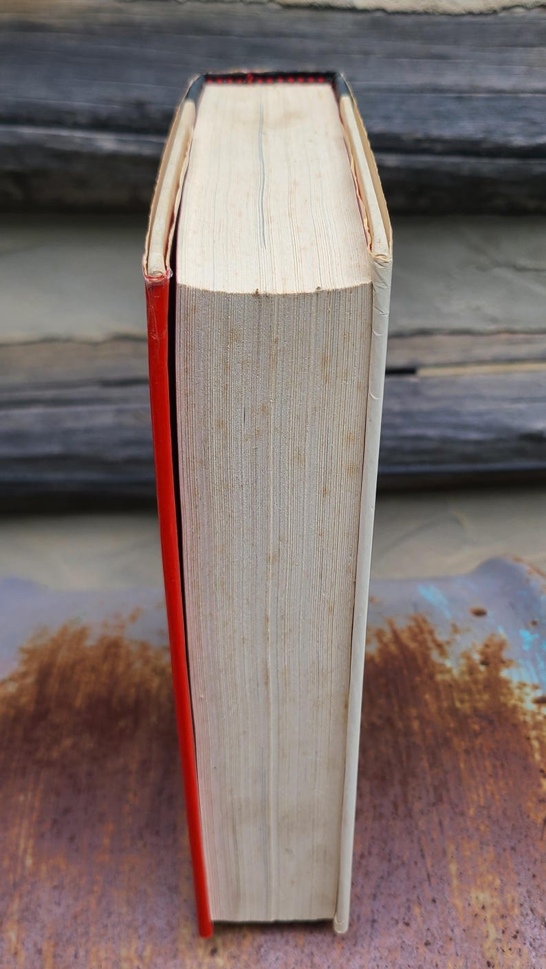 May include: A hardcover book with a red spine and white pages. The top edge of the book is visible, showing the pages and a black and red detail. The book's cover is white.