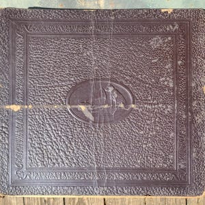 May include: A dark brown leather-bound photo album with a decorative border and a raised oval design featuring two figures. The album is slightly worn and has a vintage look.