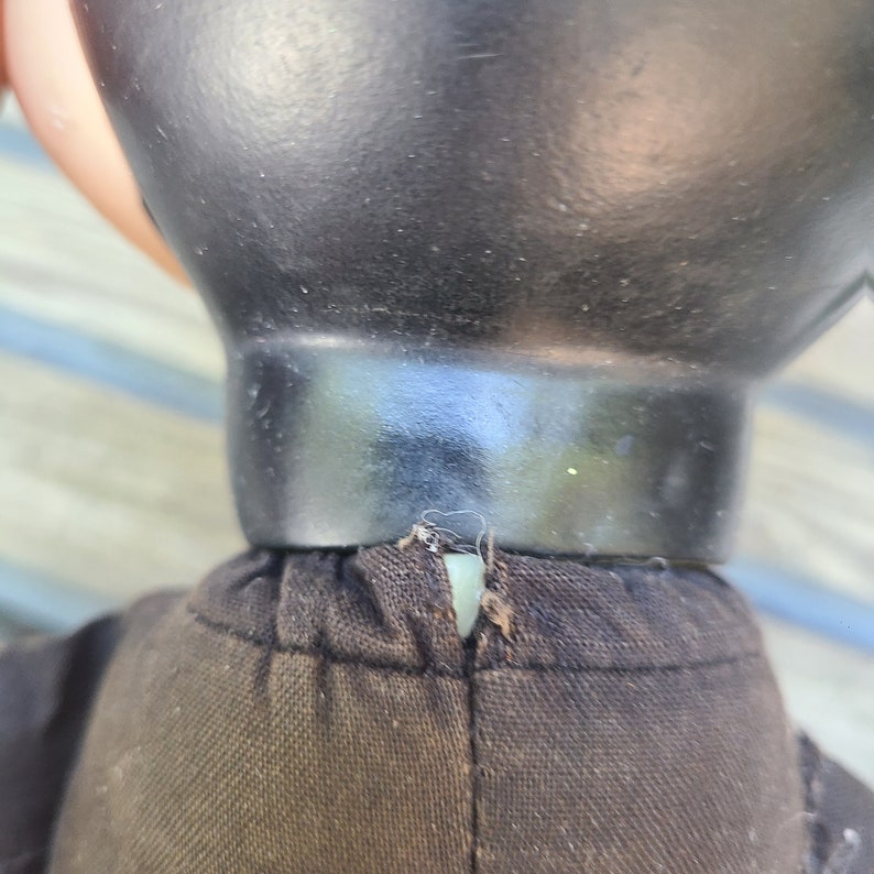May include: Close-up of the neck of a black fabric doll with a black plastic head. The doll's head is attached to the fabric body with a seam.