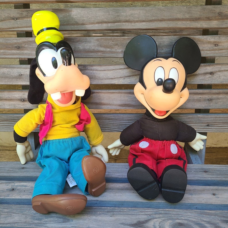 May include: Two plush toys, a yellow and brown Goofy and a black and red Mickey Mouse, are sitting on a wooden bench. Goofy is wearing a yellow shirt and blue trousers. Mickey Mouse is wearing red shorts with white polka dots.