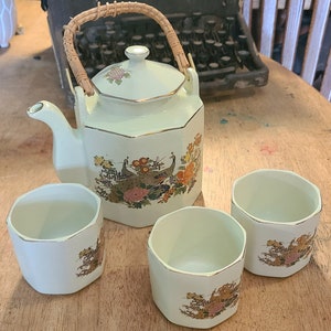 May include: A white teapot with a wicker handle and floral design, along with three matching teacups. The teapot and cups feature a gold trim and a design of peacocks and flowers.