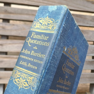 May include: A vintage book titled "Familiar Quotations" by John Bartlett. The book has a blue cover with gold lettering and decorative accents. The fourteenth edition is published by Little, Brown. The book is slightly worn, suggesting age and use.