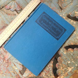 May include: A blue hardcover book titled Worship and Conduct Songs for Beginners and Primaries, positioned next to a wooden ruler that shows the book length is approximately 11 inches. The book cover features a dark blue rectangular border.