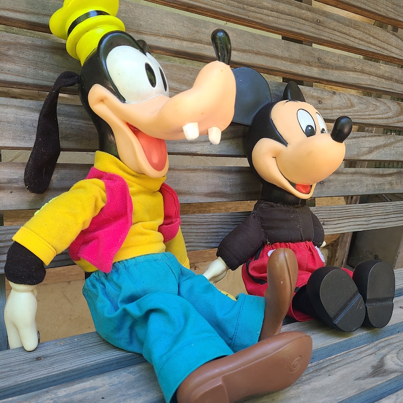 May include: A plush toy of Goofy wearing a yellow and red outfit and a blue hat, standing next to a plush toy of Mickey Mouse wearing a black and red outfit.
