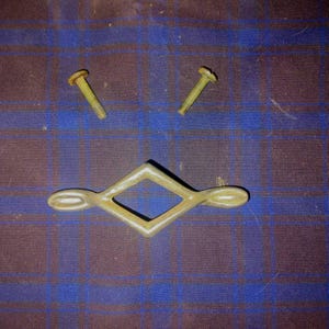 May include: A set of hardware components, including two yellow screws and a decorative metal pull with a diamond-shaped center and rounded ends. The items are arranged on a blue and brown plaid fabric.