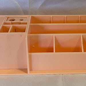 Puede incluir: Un organizador de escritorio de plástico color melocotón con múltiples compartimentos. Cuenta con un soporte para cajas de pañuelos, pequeños cubículos y varios estantes y divisores abiertos para almacenamiento. El organizador es rectangular y tiene un diseño limpio.