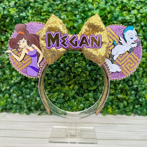 May include: Themed mouse ears with a gold sequined bow displaying the name "MEGAN" in purple. The ears are purple glitter circles with character cutouts of Megara and Pegasus. The headband is clear plastic.