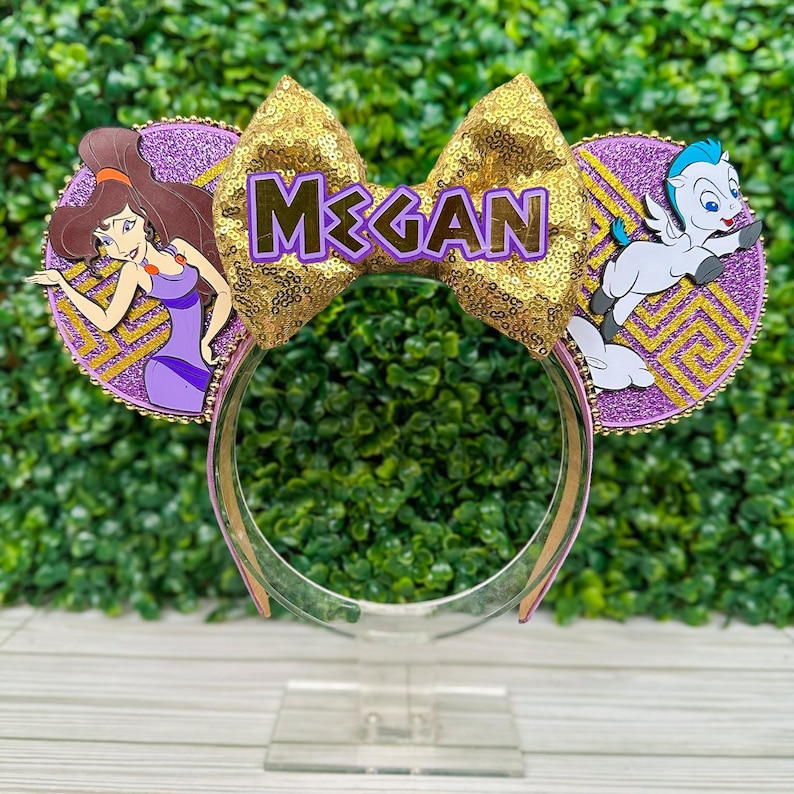 May include: A pair of themed mouse ears featuring a gold sequined bow with the name "MEGAN" in purple letters. The ears have purple glitter circles with character cutouts of Megara and Pegasus. The headband is clear.