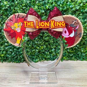May include: A themed headband featuring The Lion King. The headband has two circular ears with Simba and Pumbaa designs. A large burgundy sequined bow with a burlap centre and the words "The Lion King" completes the design.