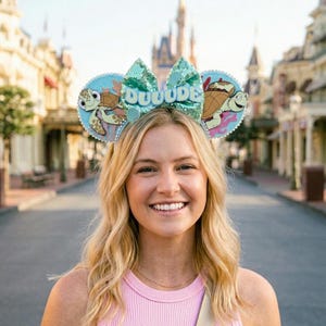 May include: A person wearing a pair of Disney-themed mouse ears. The ears feature a sequined bow with the word "DUUUDE" and cartoon turtle designs on each ear. The background shows a street with buildings.