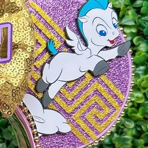 May include: A decorative headband featuring a cartoon Pegasus character with blue hair and wings, set against a purple background with gold accents. The headband has a gold sequin bow and a purple band.