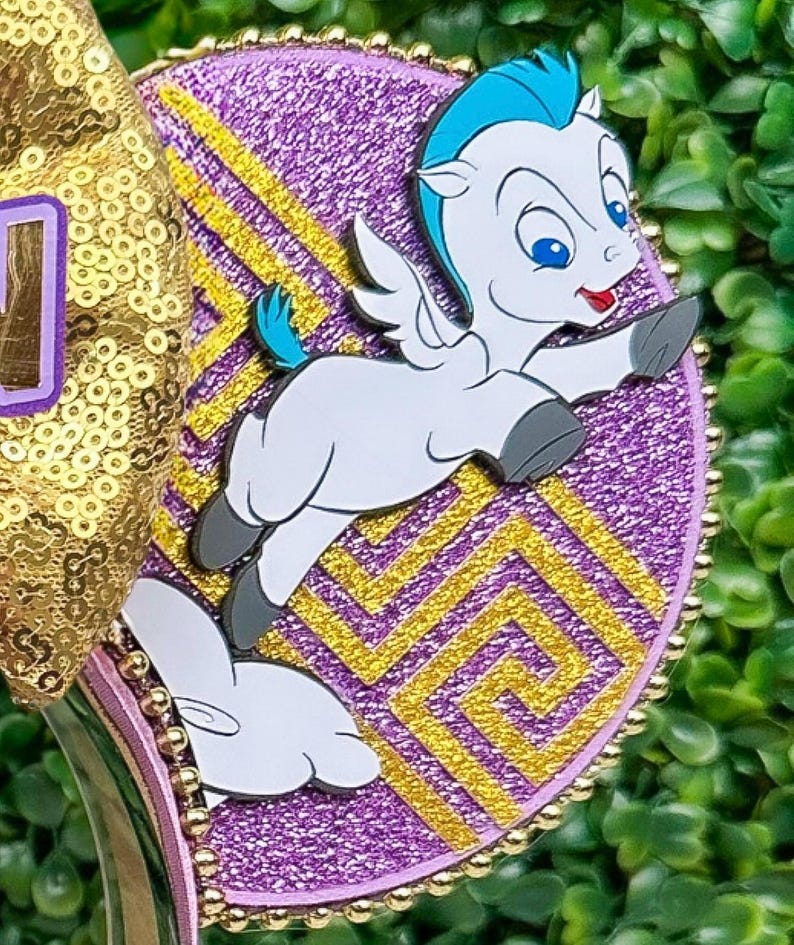 May include: A decorative headband featuring a cartoon Pegasus character with blue hair and wings, set against a purple background with gold accents. The headband has a gold sequin bow and a purple band.