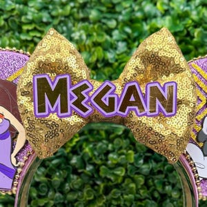 May include: A gold sequined bow with the name "Megan" in purple letters is the focal point of this headband. The headband features purple and gold accents, with cartoon character details on either side. The background is a blurred green.
