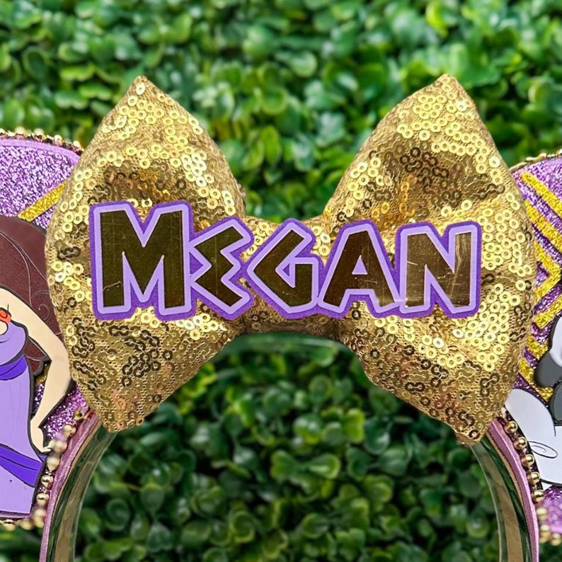 May include: A gold sequined bow with the name "Megan" in purple letters is the focal point of this headband. The headband features purple and gold accents, with cartoon character details on either side. The background is a blurred green.