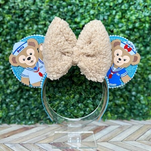 Könnte beinhalten: Ein Paar blaue und weiße Minnie Mouse Ohren mit einer braunen, flauschigen Schleife. Die Ohren zeigen ein Cartoon-Bild von Duffy und StellaLou, die als Matrosen verkleidet sind. Der Text "Duffy" und "StellaLou" befindet sich auf den Ohren.