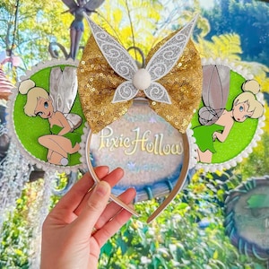 May include: Tinker Bell themed Minnie Mouse ears with a gold glitter bow, white wings, and green glitter ears featuring a Tinker Bell image. The ears are on a white headband with the text "Pixie Hollow" printed on the headband.