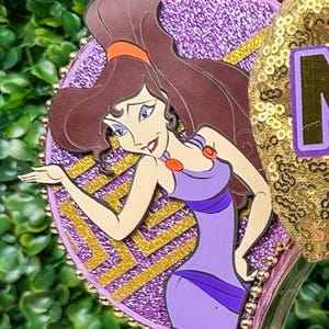 May include: A purple and gold decorative headband featuring a cartoon image of Megara from the movie Hercules. The headband has a glittery purple background with gold accents and a gold sequined ear.
