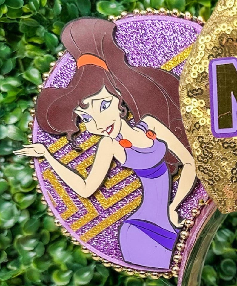 May include: A purple and gold decorative headband featuring a cartoon image of Megara from the movie Hercules. The headband has a glittery purple background with gold accents and a gold sequined ear.