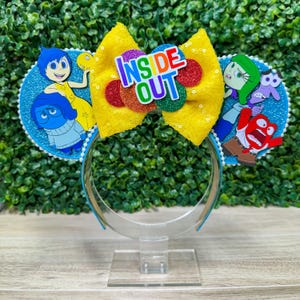 May include: A whimsical headband featuring characters from the movie "Inside Out." The headband has blue ears with character cutouts, a yellow sequined bow, and the words "Inside Out" in colorful letters. The headband is supported by a clear plastic stand.