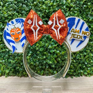 以下が含まれることがあります： 白いヘアバンドに装飾が施されています。 白いアクセントが付いたスパンコールのオレンジ色のリボンと、2つの円形の耳型のパーツがあります。 1つはキャラクターの顔、もう1つは金色の「I AM NO JEDI」と書かれています。
