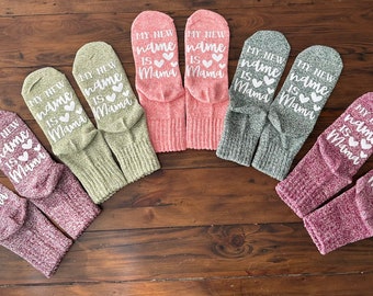May include: Six pairs of socks with a white graphic that says "My new name is Mama" with a heart symbol. The socks are in various colors including pink, green, and gray.