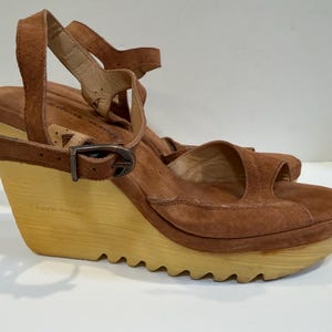 May include: A pair of brown suede wedge sandals featuring an open-toe design, an ankle strap with a metal buckle closure, and a light-colored, textured wooden wedge sole with a serrated bottom edge for added grip and a distinct profile.