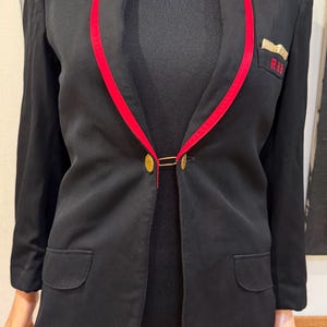 Könnte beinhalten: Schwarzer Blazer mit roter Paspel am Revers und goldenem Verschluss. Die Jacke hat zwei Vordertaschen und eine kleine Brusttasche mit rot gestickten Initialen "RH". Eine klassische, maßgeschneiderte Jacke.