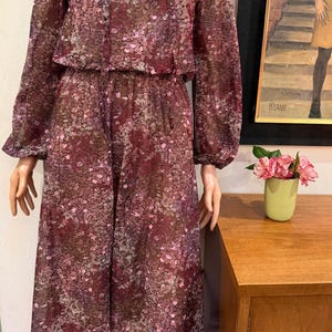 May include: A vintage floral print dress in shades of burgundy, brown, and pink. The dress features long sleeves, a V-neck, and a midi-length skirt. The dress is displayed on a mannequin. A painting and a vase of flowers are in the background.