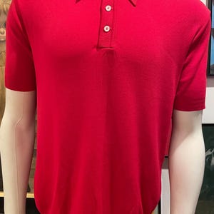 Może przedstawiać: Jasnoczerwona koszulka polo z krótkimi rękawami i klasycznym kołnierzykiem. Koszula posiada listwę z trzema guzikami i jest prezentowana na manekinie. Tkanina wydaje się być cienką dzianiną.