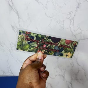 Könnte beinhalten: Ein rechteckiges Blumendesign mit dem Text "among leaves & pages I find peace" in einer kursiven Schriftart. Das Design zeigt grüne Blätter, Beeren und Blumen. Der Hintergrund ist ein weißes Marmormuster.