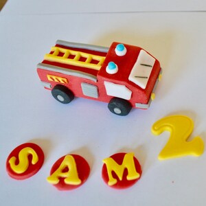 May include: Red and gray fire truck cake topper with a yellow ladder and blue lights. The topper is surrounded by red circles with yellow letters spelling out "SAM" and a yellow number "2".