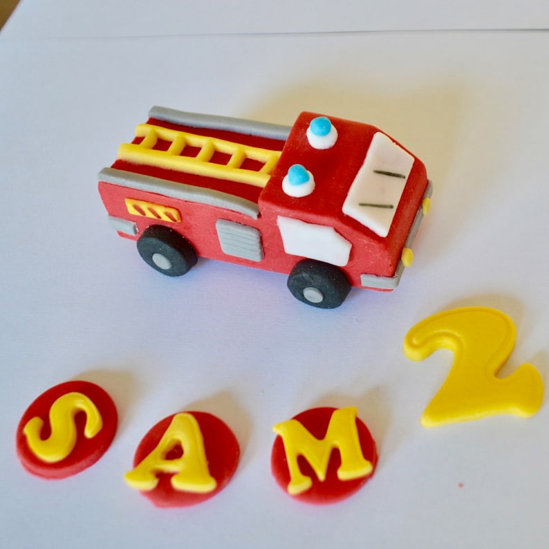 May include: Red and gray fire truck cake topper with a yellow ladder and blue lights. The topper is surrounded by red circles with yellow letters spelling out "SAM" and a yellow number "2".