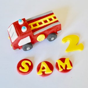 May include: Red and white fire truck cake topper with a yellow ladder and black wheels. The topper is surrounded by yellow letters spelling "SAM" and the number "2" in yellow.