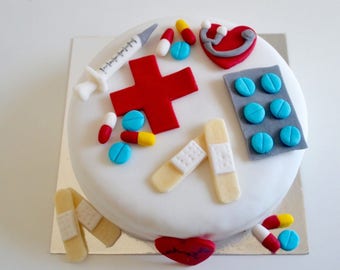 May include: A white cake decorated with a red cross, blue pills, bandages, a red heart, and a syringe. The cake is on a silver cake stand.