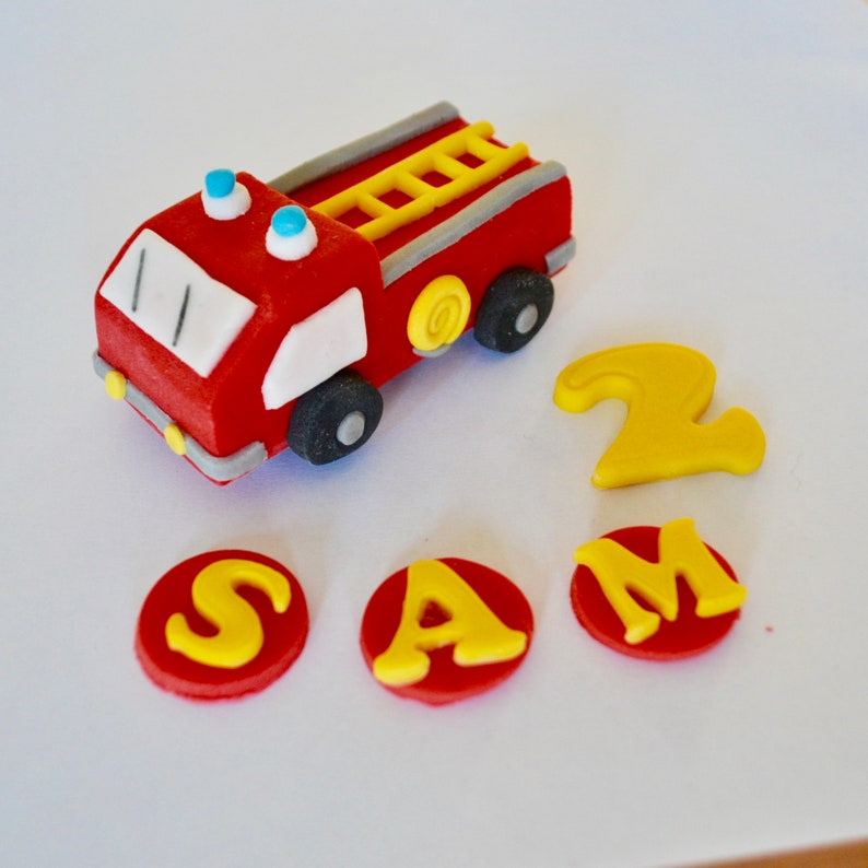 May include: Red and white fire truck cake topper with a yellow ladder and blue lights. The topper is surrounded by red circles with yellow letters spelling out "SAM 2".