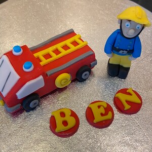 May include: A red fire truck cake topper with a yellow ladder and blue lights. A yellow and blue firefighter figurine stands next to the truck. The letters B, E, and N are on the cake in yellow letters on red circles.