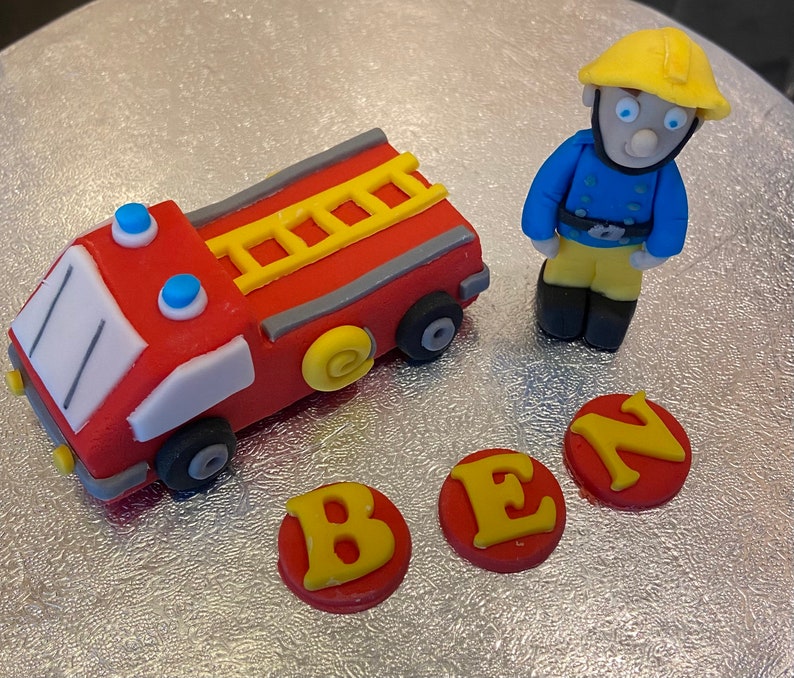 May include: A red fire truck cake topper with a yellow ladder and blue lights. A yellow and blue firefighter figurine stands next to the truck. The letters B, E, and N are on the cake in yellow letters on red circles.